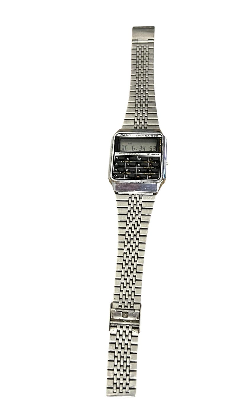 May include: Silver-toned Casio CA-502 calculator watch. The watch features a rectangular digital display showing the time, with a full calculator keypad below. The watch band is made of small, connected metal links.