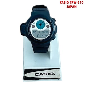 May include: A black Casio CPW-310 digital watch with a white face displaying the time and date. The watch has a black strap and is displayed on a white stand. The text "CASIO" is visible on the watch face and the stand.
