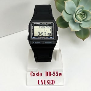 Puede incluir: Un reloj digital Casio DB-55W negro con una esfera rectangular que muestra la hora y la fecha. El reloj tiene una correa negra y el texto "Casio DB-55w UNUSED" es visible debajo.