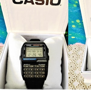 Puede incluir: Un reloj digital Casio Data Bank 80 negro con una calculadora y una pantalla plateada. El reloj tiene una correa negra y una esfera plateada. La pantalla muestra la fecha, la hora y el día de la semana. El reloj está en una caja blanca.