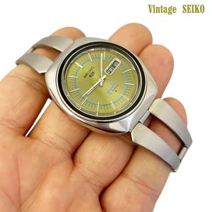 May include: Vintage Seiko watch with a silver-tone case and bracelet. The watch face is olive green with blue accents and displays the day and date. The watch is labeled "SPORT 21 JEWELS" and "FRI 18". The text "Vintage SEIKO" is in the upper right corner.