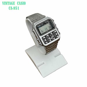 May include: A silver-tone Casio CA-951 digital watch with a metallic link strap. The watch face features a rectangular LCD screen displaying the time and a built-in calculator keypad with white buttons labelled with numbers and symbols.