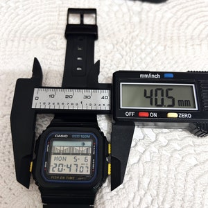 May include: A black Casio digital watch with a yellow trim and a blue display. The watch face shows the date, time, and a tide graph. The watch is being measured with a digital caliper, which reads 40.5 mm.