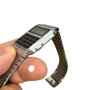 May include: Silver-toned digital watch with calculator keys and a mesh band. The watch face displays a digital screen and the text "CASIO CA-502". A vintage-style accessory.