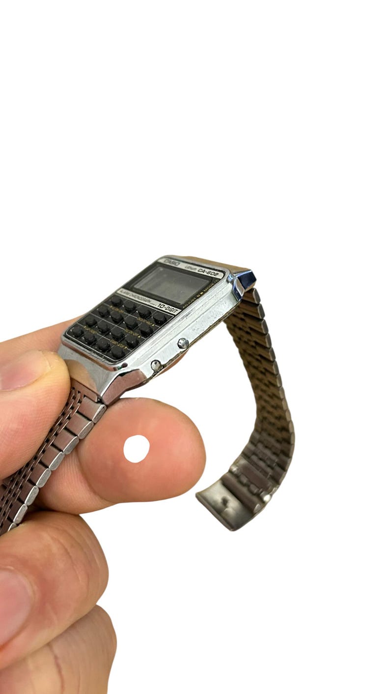 May include: Silver-toned digital watch with calculator keys and a mesh band. The watch face displays a digital screen and the text "CASIO CA-502". A vintage-style accessory.