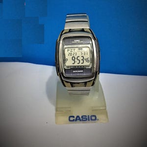 Puede incluir: Un reloj Casio Tough Solar plateado con una esfera negra y una pantalla digital blanca. El reloj está en un soporte blanco con el logotipo de Casio.