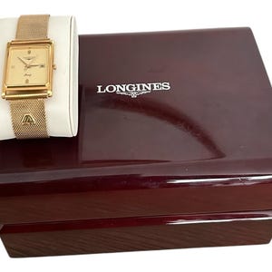 May include: A gold-toned Longines watch with a rectangular face and mesh band rests on a white cushion. The watch is displayed next to a dark burgundy wooden box with the Longines logo in white.