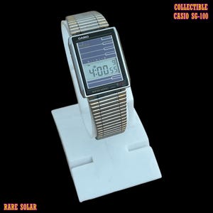 Pode incluir: Um relógio digital Casio SG-100 prateado com mostrador retangular, pulseira de elos metálicos e painel solar. O ecrã mostra a hora 4:00:55. O texto no mostrador inclui Casio e Water Resist. O texto na imagem diz Collectible Casio SG-100 Rare Solar.