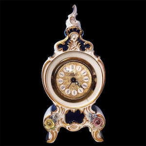 May include: A white porcelain clock with gold accents and a blue and white base. The clock has Roman numeral markings and a gold-toned face.