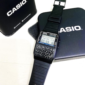 Puede incluir: Reloj digital Casio DBX-102 negro con teclado de calculadora y correa negra. El reloj muestra la hora y la fecha. El reloj está encima de una caja negra con el logotipo de Casio.
