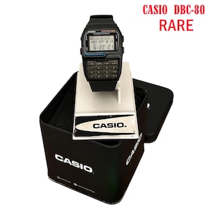 Puede incluir: Un reloj Casio DBC-80 negro con teclado de calculadora y pantalla digital, exhibido sobre un soporte blanco dentro de una caja negra. El reloj muestra la fecha y la hora. La caja tiene el logotipo de Casio.