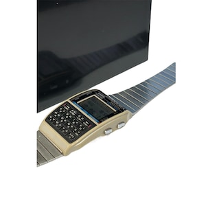 May include: Vintage Casio calculator watch with a beige case, black keyboard, and a silver-tone metal band. The watch features a digital display and several buttons. The watch is displayed against a black background.
