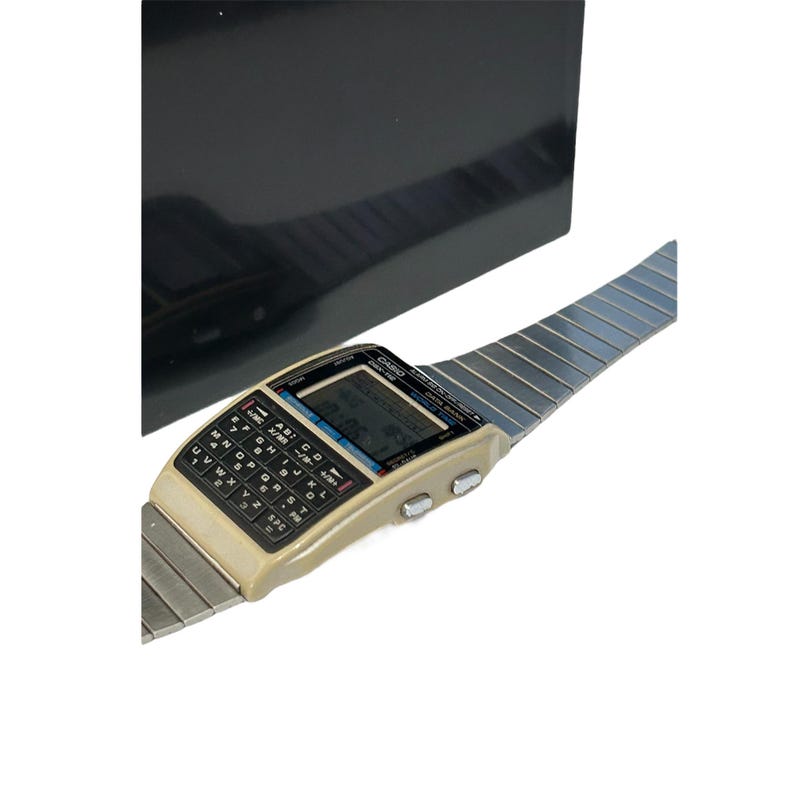 May include: Vintage Casio calculator watch with a beige case, black keyboard, and a silver-tone metal band. The watch features a digital display and several buttons. The watch is displayed against a black background.