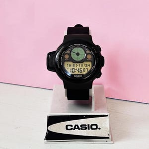 May include: Black Casio digital watch with a multi-function display. The watch face shows the time, date, and day of the week. The watch is on a black band and is sitting on a white display stand with the Casio logo.