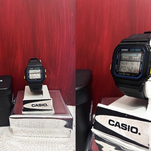 May include: A black Casio digital watch with a yellow and blue trim. The watch is on a white display stand and has a black box with the Casio logo.