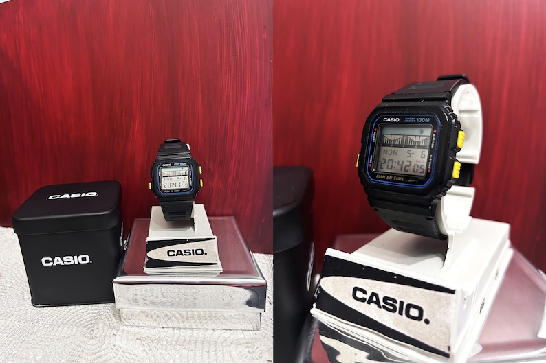 May include: A black Casio digital watch with a yellow and blue trim. The watch is on a white display stand and has a black box with the Casio logo.