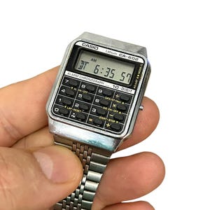 May include: A vintage Casio CA-602 digital watch with a silver-tone case and band. The watch face displays the time as 6:35:57 AM. Features include an alarm, chronograph, and 10-digit calculator functions. The watch is held against a white background.