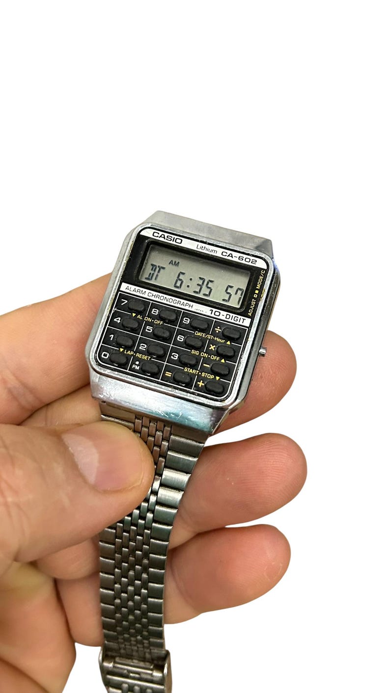 May include: A vintage Casio CA-602 digital watch with a silver-tone case and band. The watch face displays the time as 6:35:57 AM. Features include an alarm, chronograph, and 10-digit calculator functions. The watch is held against a white background.