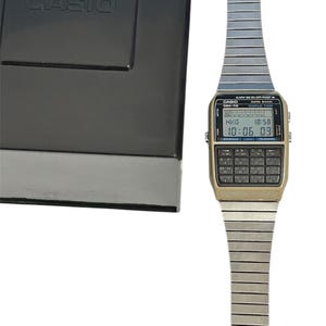 May include: A vintage Casio calculator watch with a silver-tone band and a gold-tone face. The watch displays the time and has a calculator keypad. The watch is next to a black box with the Casio logo.