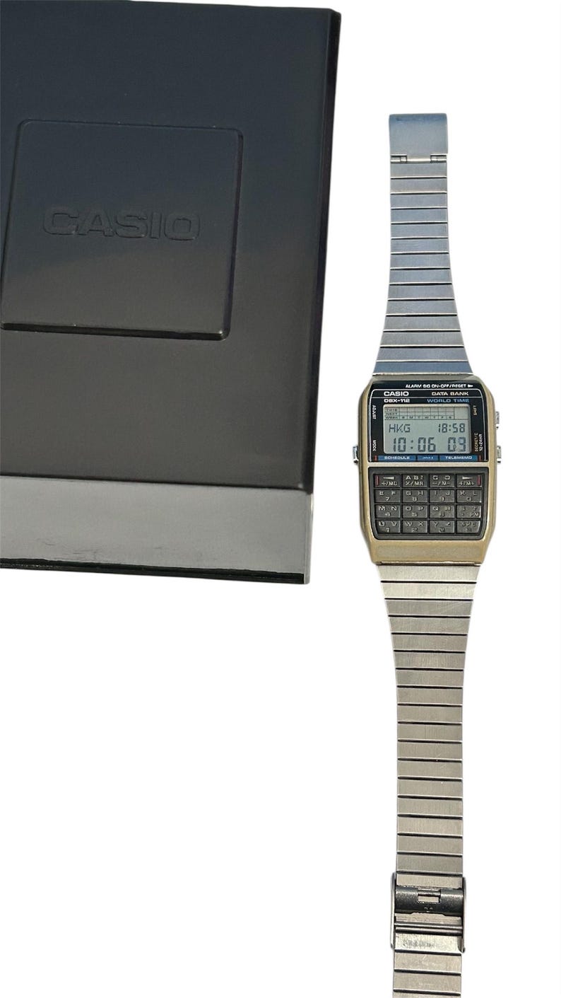 May include: A vintage Casio calculator watch with a silver-tone band and a gold-tone face. The watch displays the time and has a calculator keypad. The watch is next to a black box with the Casio logo.