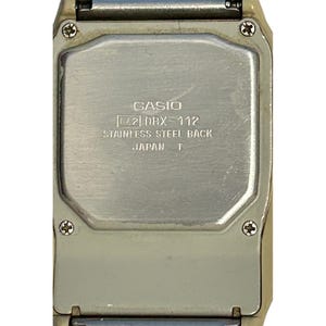 May include: Close-up of the back of a Casio DBX-112 digital watch. The watch has a stainless steel back and is marked "Japan T". The watch case is a light beige color.