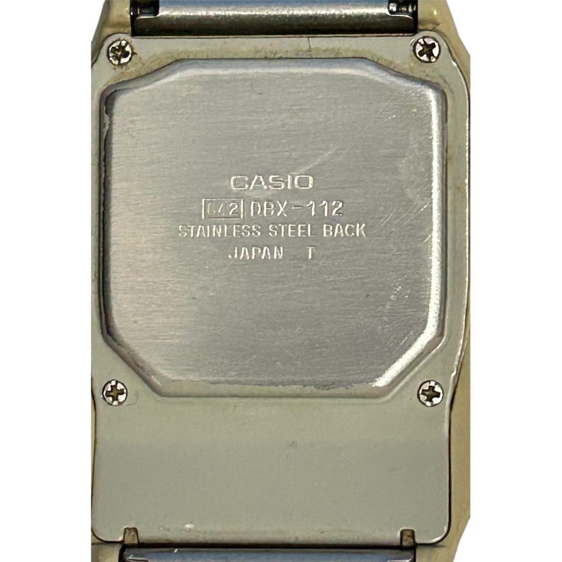 May include: Close-up of the back of a Casio DBX-112 digital watch. The watch has a stainless steel back and is marked "Japan T". The watch case is a light beige color.