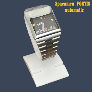 Op de afbeelding: Een zilveren Spacemen Fortis automatische horloge met een zwarte vierkante wijzerplaat. Het horloge heeft een zilveren band en wordt gepresenteerd op een witte standaard. De wijzerplaat heeft gouden markeringen en rode en zilveren wijzers. De tekst "Spacemen Fortis automatic" is geel.