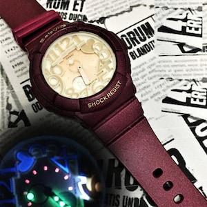 May include: A burgundy and white Casio Baby-G watch with a shock resistant feature. The watch face has a unique design with large numbers and a heart shape.