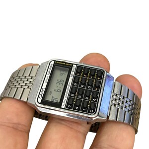 May include: A silver Casio CA-602 digital watch with a calculator keypad. The watch displays the time as 6:36:43 AM. The watch has a silver metal band and is held in a hand.
