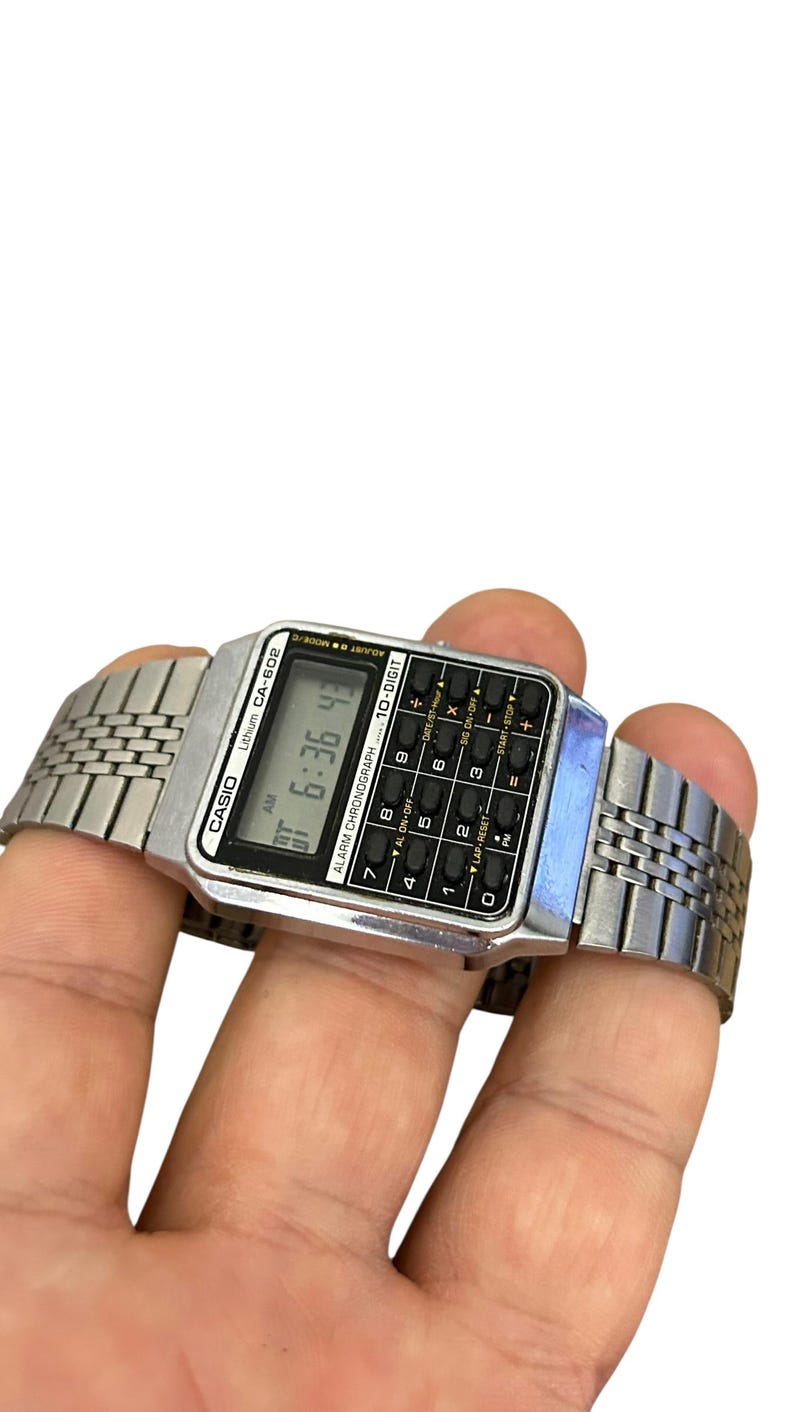 May include: A silver Casio CA-602 digital watch with a calculator keypad. The watch displays the time as 6:36:43 AM. The watch has a silver metal band and is held in a hand.