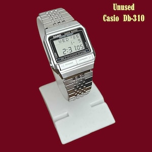 Può includere: Un orologio digitale Casio DB-310 argentato su un espositore bianco. Il quadrante mostra la data, l'ora e il giorno della settimana. Il cinturino è realizzato con maglie in metallo argentato. Il testo "Unused Casio Db-310" è in alto a destra.