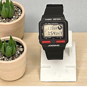 Puede incluir: Reloj digital Casio negro con esfera rectangular, botones rojos y la palabra "JOGGING" impresa en la parte inferior. El reloj muestra la hora y otros datos. El reloj está sobre un soporte blanco. Dos pequeñas suculentas en macetas están en el fondo.
