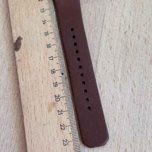 May include: A brown calculator watch with a brown strap. The watch is next to a wooden ruler. The ruler shows measurements from 10 to 25 inches.