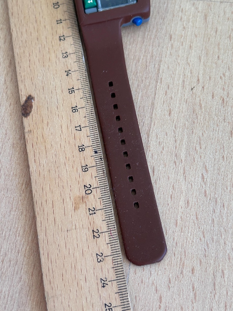May include: A brown calculator watch with a brown strap. The watch is next to a wooden ruler. The ruler shows measurements from 10 to 25 inches.