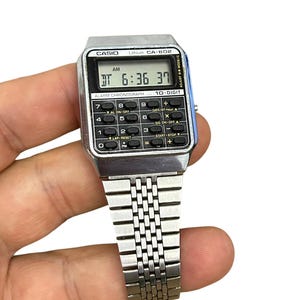May include: A silver Casio CA-602 digital watch with a calculator function. The watch displays the time as 6:36:37 AM. It has a silver band and a black digital display with buttons for various functions.