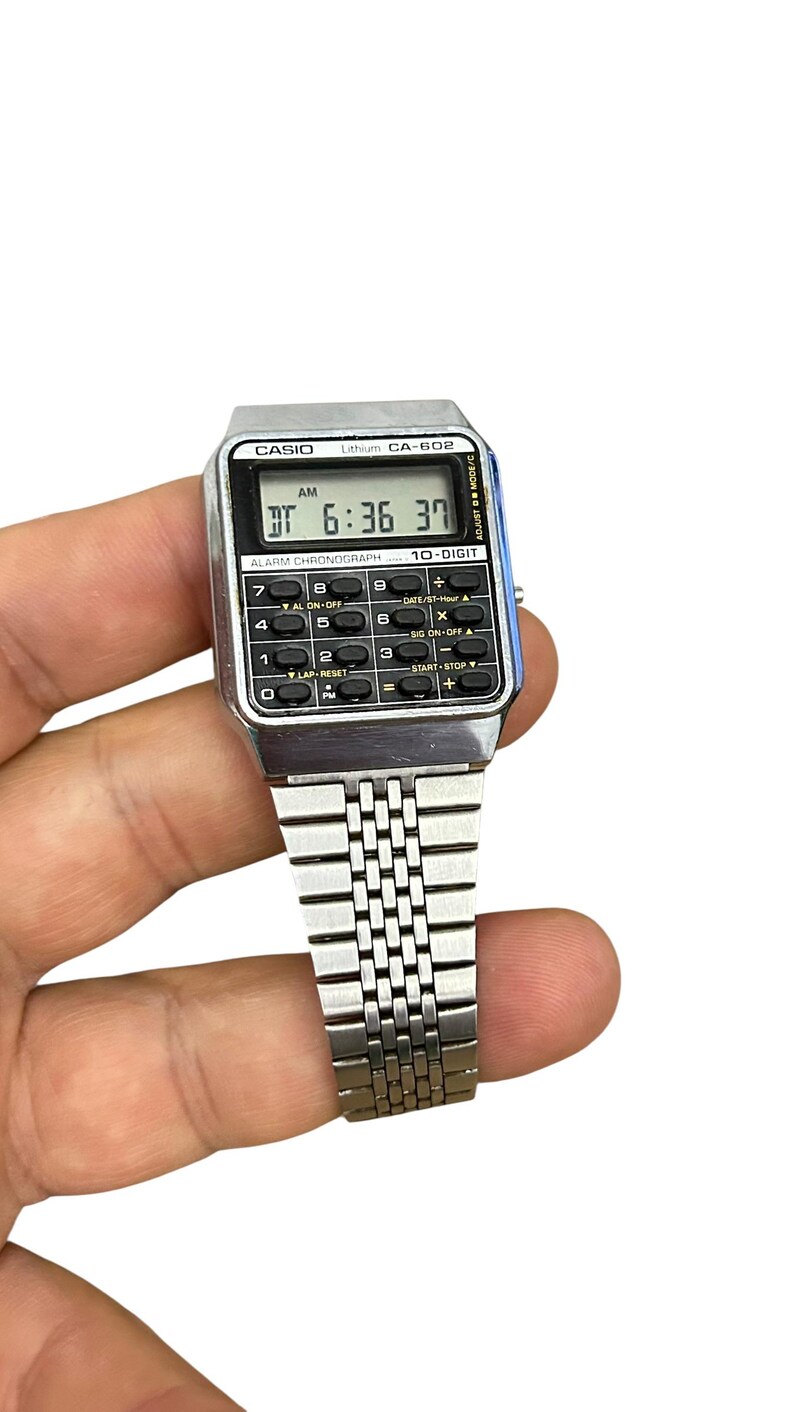 May include: A silver Casio CA-602 digital watch with a calculator function. The watch displays the time as 6:36:37 AM. It has a silver band and a black digital display with buttons for various functions.