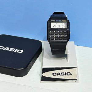 May include: A black Casio calculator watch with a digital display showing the time 2:09 and the date TH 04. The watch has a black band and a black case. The watch is on a white display stand with the word "CASIO" printed on it.