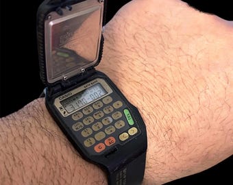 Könnte beinhalten: Eine schwarze Casio Data Bank Taschenrechneruhr mit silbernem Zifferblatt und einer aufklappbaren Abdeckung. Das Zifferblatt der Uhr zeigt Datum und Uhrzeit an, und der Taschenrechner verfügt über verschiedene Funktionen, darunter Addition, Subtraktion, Multiplikation, Division und Prozentrechnung.