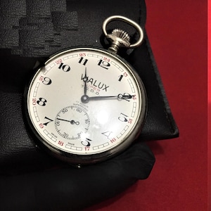 May include: A silver pocket watch with a white face and black hands. The watch has a black leather case and the words "WALUX SUPER T.C.D.D." printed on the face.