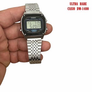 May include: A silver Casio DW-1400 digital watch with a black face displaying the date and time. The watch has a stainless steel band and is held in a hand. Text on the watch face reads "CASIO". The text "ULTRA RARE CASIO DW-1400" is in the upper right corner.