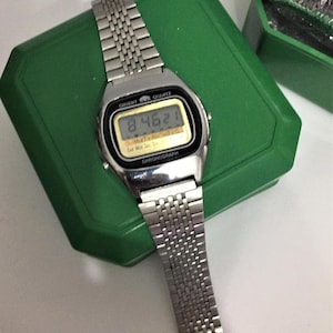 Könnte beinhalten: Eine silberne Orient Chronograph-Armbanduhr mit einem schwarzen digitalen Display, das die Zeit 8:46:21 anzeigt. Die Uhr hat ein silbernes Metallband und befindet sich in einer grünen Box.