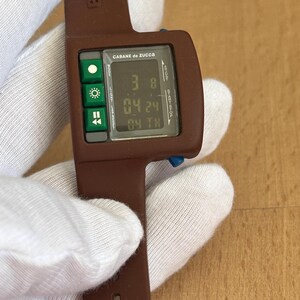 May include: A brown digital wristwatch with a rectangular face and a matching band. The watch face displays the time and date, with green buttons on the left side. The brand name "CABANE de ZUCCA" is printed on the watch face.