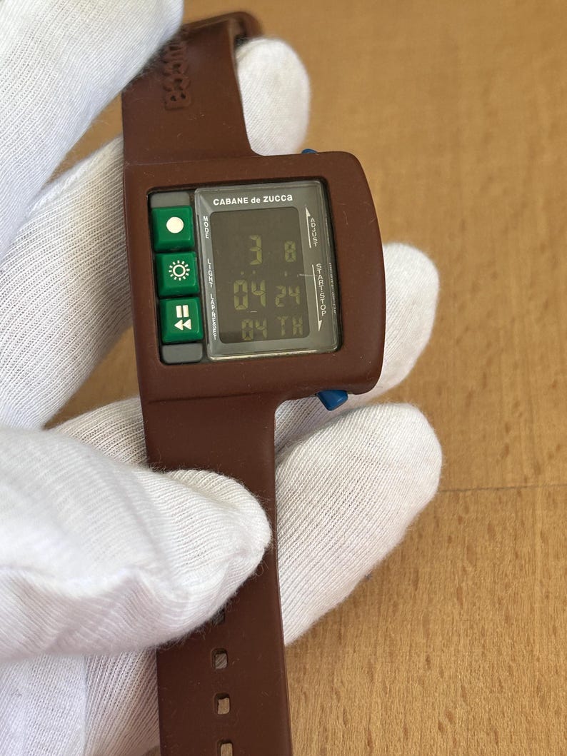 May include: A brown digital wristwatch with a rectangular face and a matching band. The watch face displays the time and date, with green buttons on the left side. The brand name "CABANE de ZUCCA" is printed on the watch face.