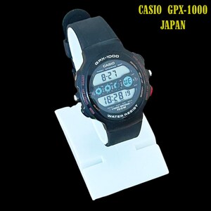 May include: A black Casio GPX-1000 digital watch with a black band, displayed on a white stand. The watch face shows the time, date, and other functions. The text "CASIO GPX-1000 JAPAN" is visible in the upper right corner.