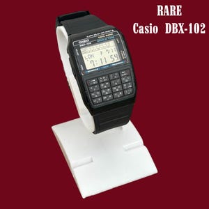 Puede incluir: Un reloj digital Casio DBX-102 negro con un teclado de calculadora y una pantalla digital que muestra la hora. El reloj está sobre un soporte de exhibición blanco sobre un fondo granate. El texto "RARE" y "Casio DBX-102" son visibles.