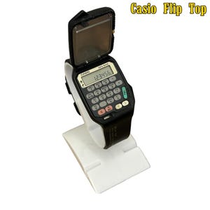 Può includere: Un orologio nero Casio Flip Top con un quadrante calcolatrice, che mostra il numero 123456. L'orologio ha una copertura a ribalta ed è esposto su un supporto bianco. Il testo "Casio Flip Top" è visibile.