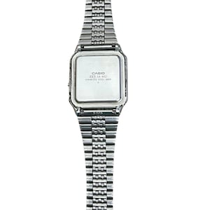 May include: A silver-toned Casio digital watch with a rectangular face and a stainless steel band. The watch face has the text "CASIO" and "CA-602". The band is made of small, connected metal links.