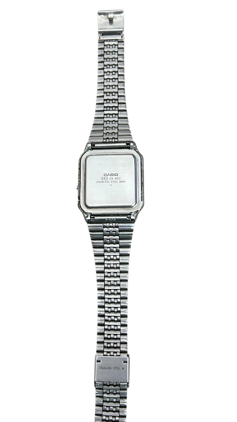 May include: A silver-toned Casio digital watch with a rectangular face and a stainless steel band. The watch face has the text "CASIO" and "CA-602". The band is made of small, connected metal links.
