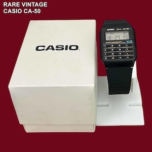 Puede incluir: Un reloj digital Casio CA-50 vintage con una correa negra y botones estilo calculadora, exhibido en un soporte blanco. La esfera del reloj muestra la hora. El reloj está junto a su caja blanca original con el logotipo de Casio.
