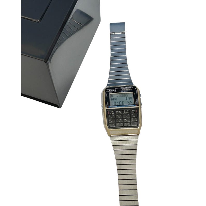 May include: A vintage Casio calculator watch with a gold-tone face and a silver-tone metal band. The watch displays the time and date on a digital screen and features a full keypad. The watch is next to a black, glossy box.